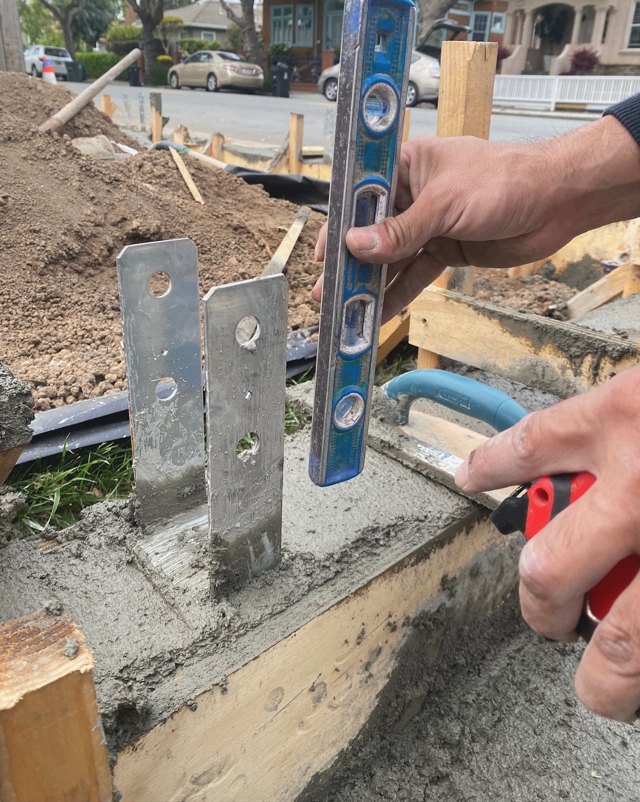 Hands holding a spirit level against galvanised post bases set into a freshly poured concrete footing, with drain rock and filter fabric behind.