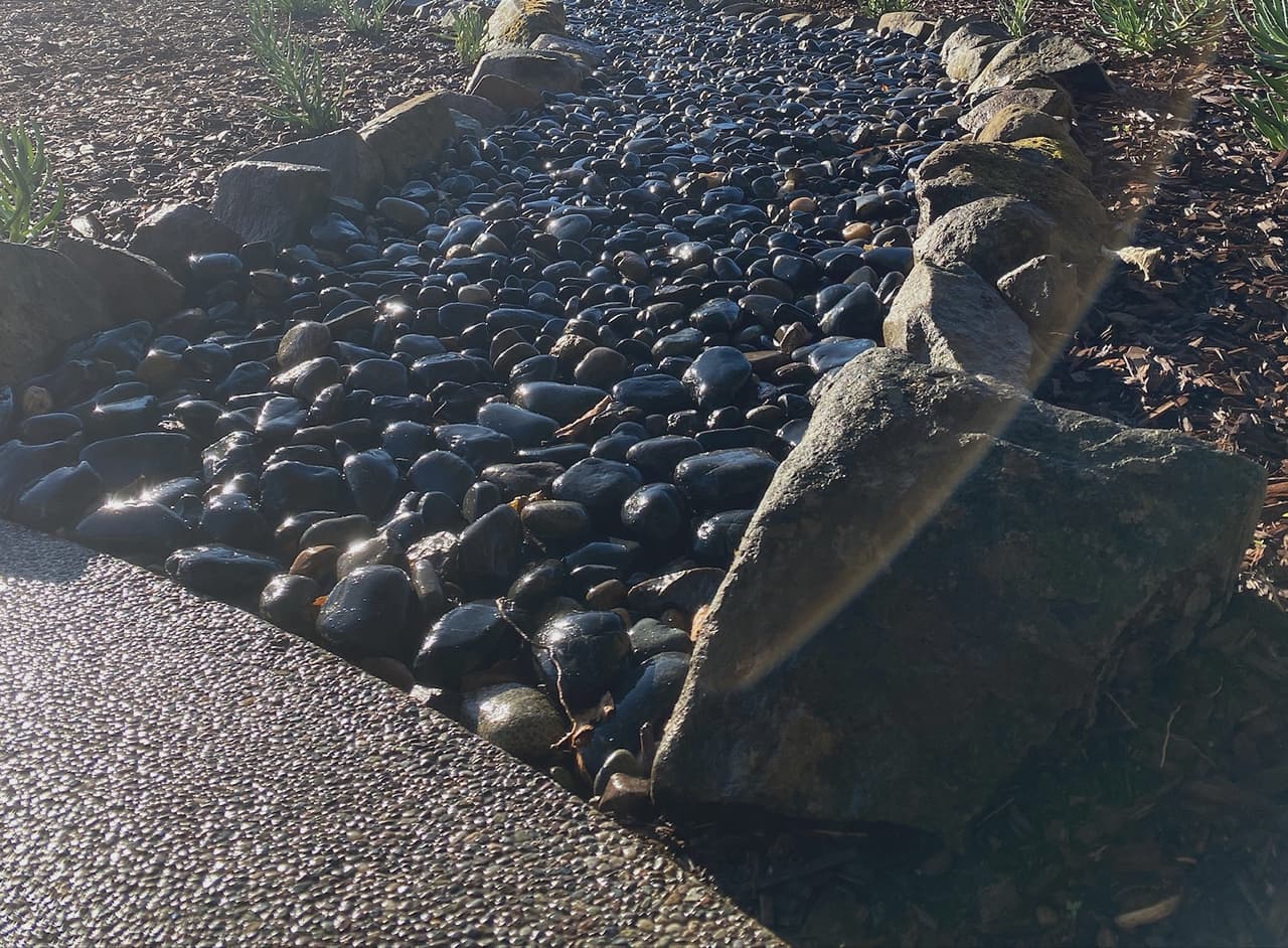 A dry creek bed of dark river rock between boulder banks, carrying runoff across a planted front garden.