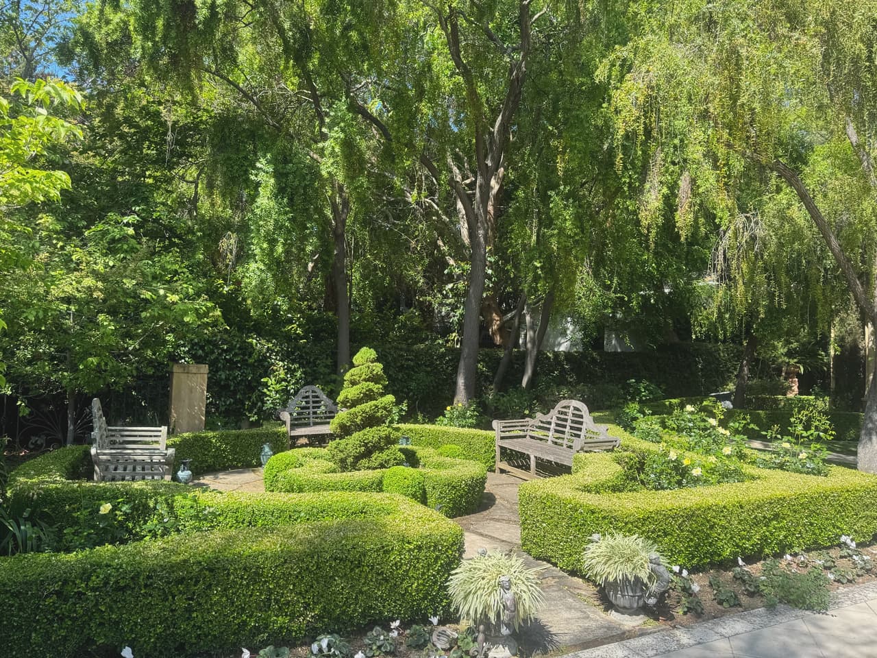 A formal boxwood knot garden with a clipped spiral topiary at its centre and teak benches set into the hedge bays.