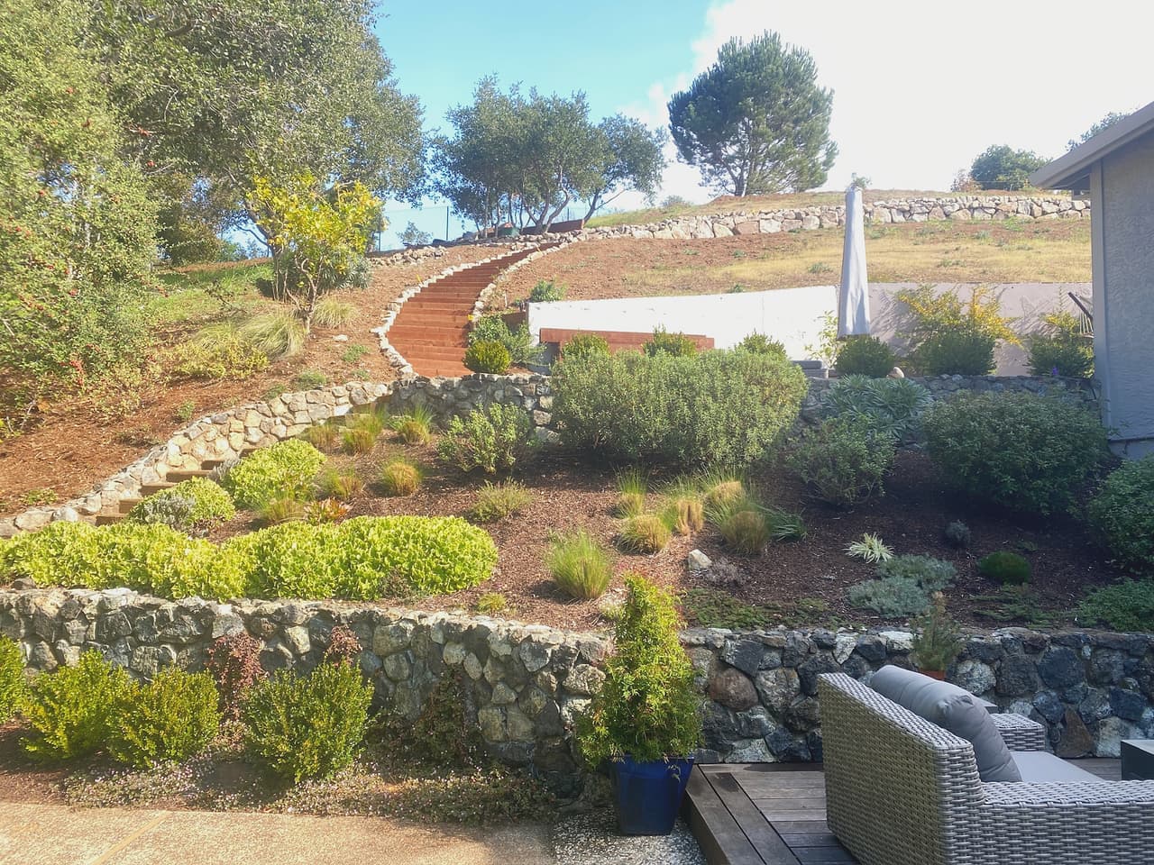 A hillside terraced with dry-laid cobble retaining walls, each bench planted with low shrubs above a timber deck.